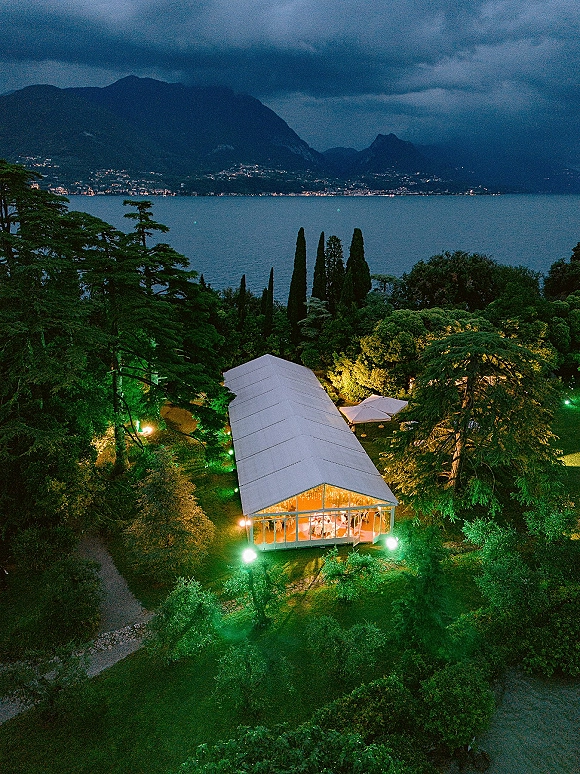 Outdoor reception tent with cafe lights glowing under a cloudy mountain sky beside a lake, with uplighting in trees and umbrellas