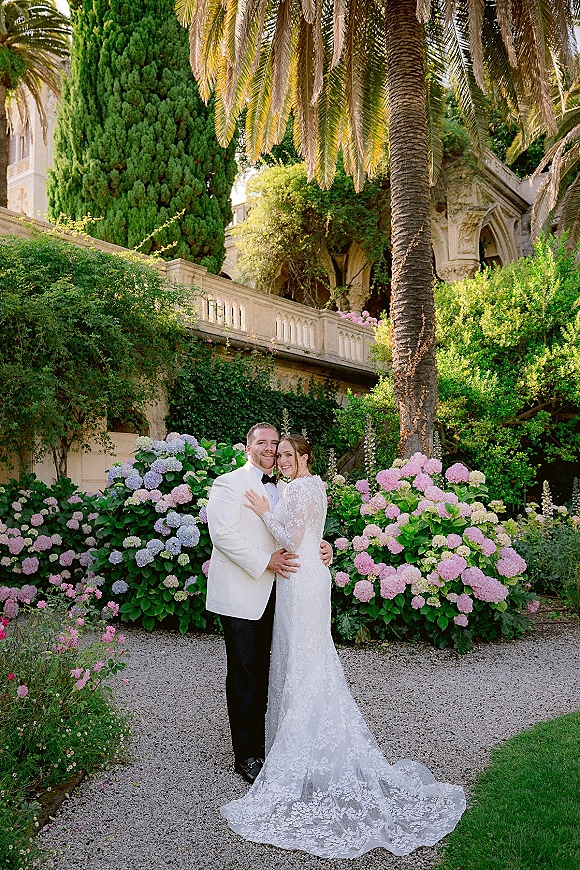 Couple portrait of bride and groom embrace, smiling on a stone terrace garden with palm trees, ivy, and hydrangeas backdrop