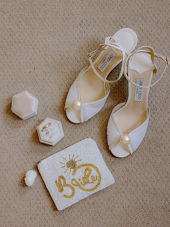 Bridal accessories flatlay with white ankle-strap heels, pearl embellishments, hexagon ring box, rings, and beaded clutch on beige carpet