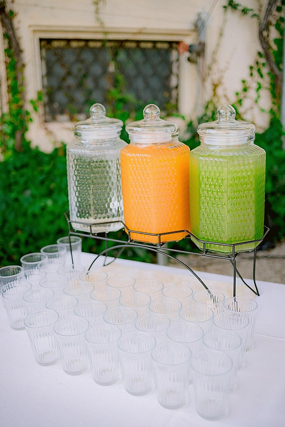 Wedding drink station with glass beverage dispensers on a metal stand, plastic cups on white tablecloth, set against ivy and a window
