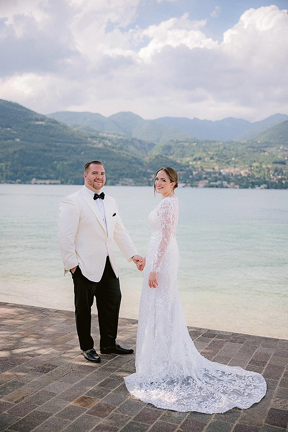 Couple portrait of bride and groom holding hands, her long sleeve lace gown and his white jacket on a stone terrace by lake and mountains