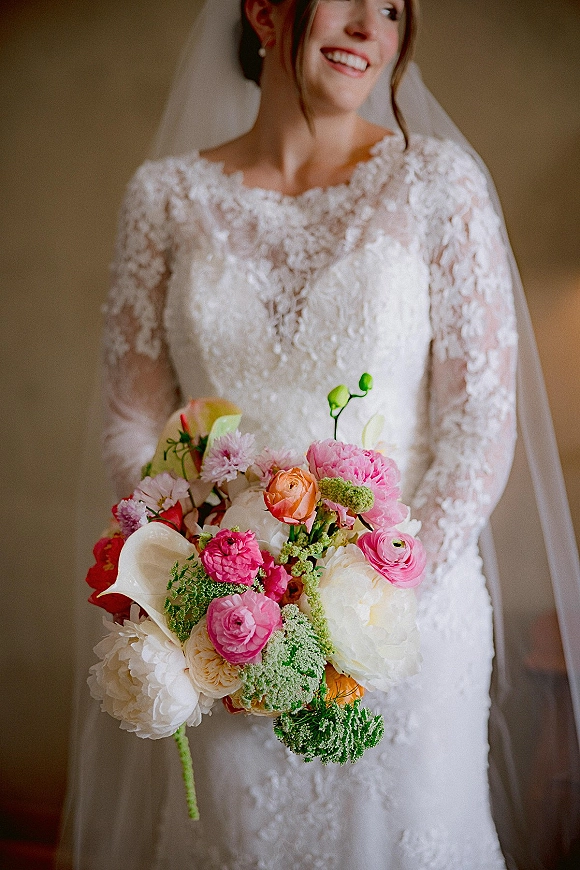 Bridal portrait of a smiling bride holding bouquet in a long-sleeve lace gown and veil, with pearl drop earrings by a neutral wall indoors