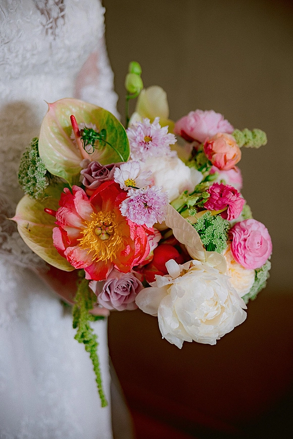 Bridal bouquet with colorful wedding bouquet blooms—peonies, roses, anthurium, ranunculus and greenery—held against a lace dress by a neutral wall