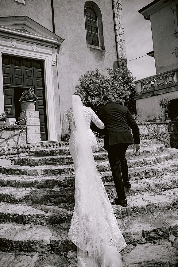 Wedding couple portrait of bride and groom holding hands walking up stone church steps, her long veil and lace train flowing behind