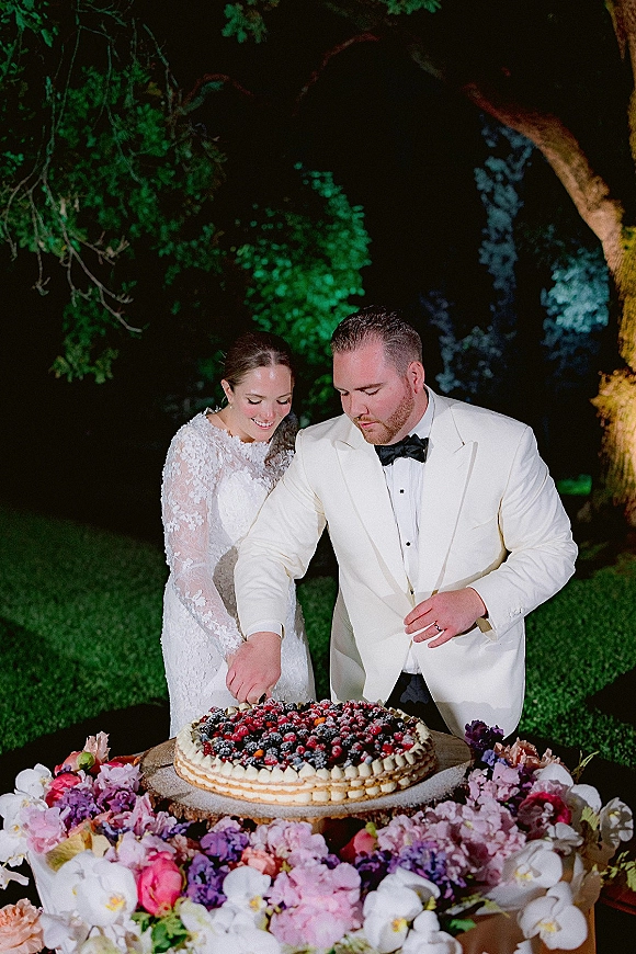 Wedding cake cutting as bride in long sleeve lace dress and groom in white tuxedo slice berry-topped cake outdoors at night with orchids