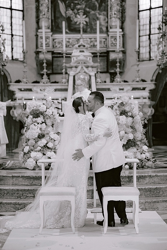 Ceremony kiss as bride in lace sleeves and cathedral veil kisses groom in tuxedo at church altar with stained glass windows behind