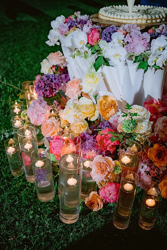 Wedding cake display with a white floral-topped cake, surrounded by roses, orchids, hydrangeas and candlelit glass vases on a grassy lawn at night