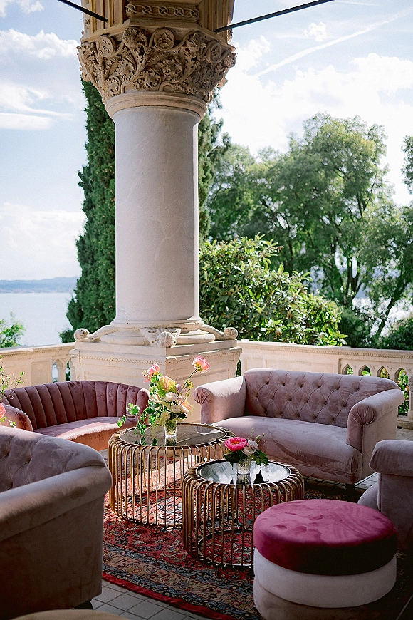 Lounge seating area with wedding lounge seating on blush velvet sofas, round glass coffee tables, floral centerpieces, on a stone terrace by a lake view