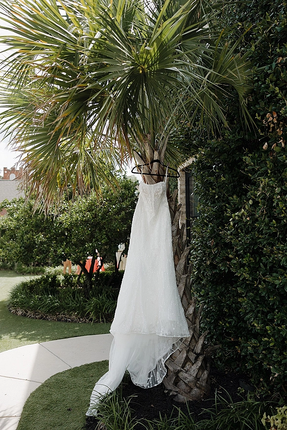 Wedding dress with lace details hanging on a hanger, the train draping in front of a palm tree and green garden hedge outdoors