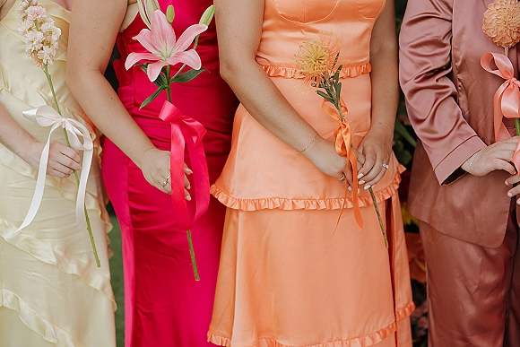 Bridesmaid dresses in a mismatched bright palette, each holding single-stem flowers with satin ribbon bows amid lush garden greenery