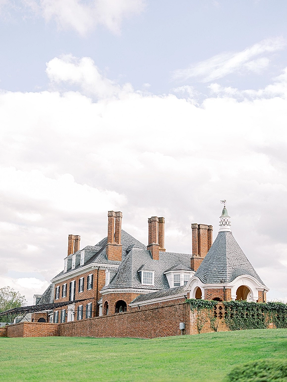 Wedding venue exterior of a brick estate wedding venue with ivy, turret, chimneys and string lights, set on a green lawn under cloudy sky
