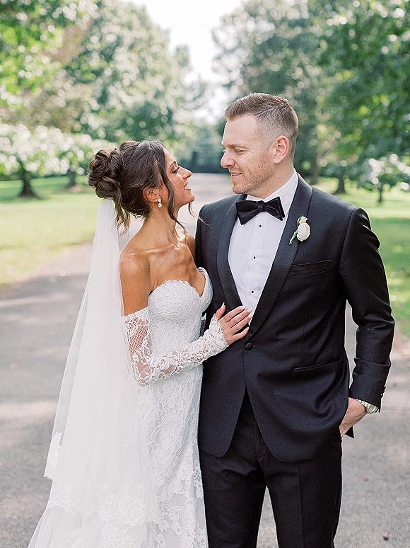 Couple portrait of bride looking at groom, holding his lapel, her veil trailing, on a tree-lined path with daylight greenery