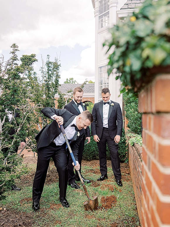 Groomsmen photo of men in black tuxedos and bow ties posing with a shovel on a lawn by a brick wall and shrubs under clouds