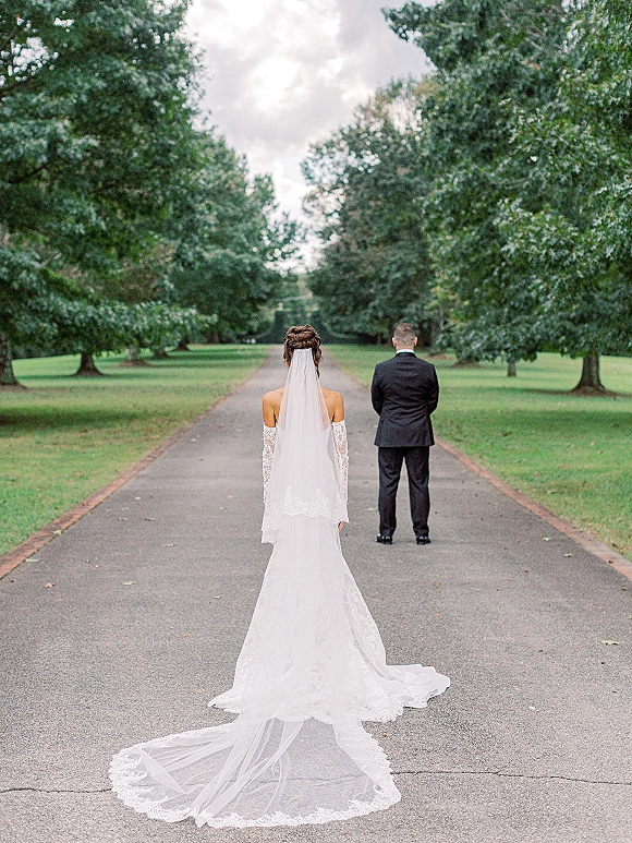 Wedding first look as bride in lace dress with long veil approaches groom in suit on a tree-lined road under cloudy sky