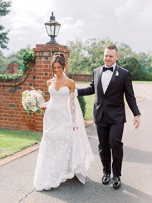 Couple portrait of bride and groom walking together, bride holding bouquet, beside a brick wall with lantern and greenery outdoors