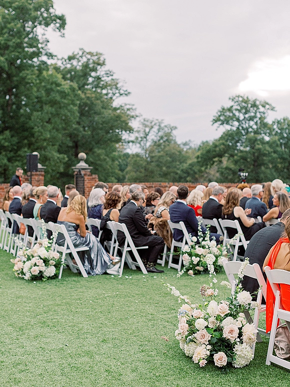 Outdoor wedding ceremony with rows of wedding ceremony seating, white folding chairs flanking a rose-and-hydrangea floral aisle on a lawn