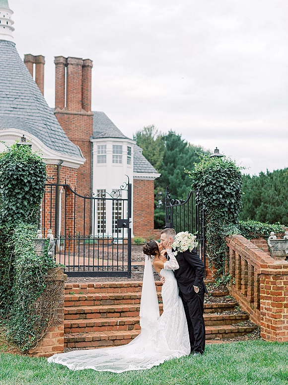 Wedding kiss portrait of bride and groom kissing, bride holding white rose bouquet with veil on brick steps by ivy and wrought iron gate