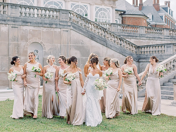 Bridal party portrait of the bride with bridesmaids holding bouquets on a stone staircase, lace sleeves and neutral dresses by arched windows