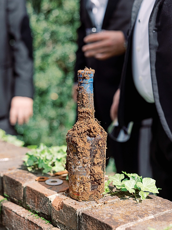 Wine bottle detail with dirt and ivy vines resting on a brick ledge, blurred wedding guests in suits in the greenery behind