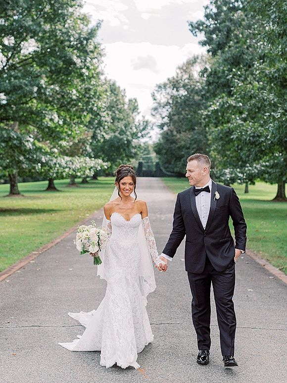 Couple portrait of bride and groom walking hand in hand, bride in off-shoulder lace dress holding bouquet on tree-lined driveway