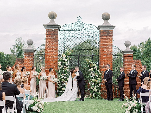 Ceremony kiss as the bride and groom kiss beneath a blush-and-white floral arch with greenery, bridal party applauding by brick pillars and a wrought iron gate