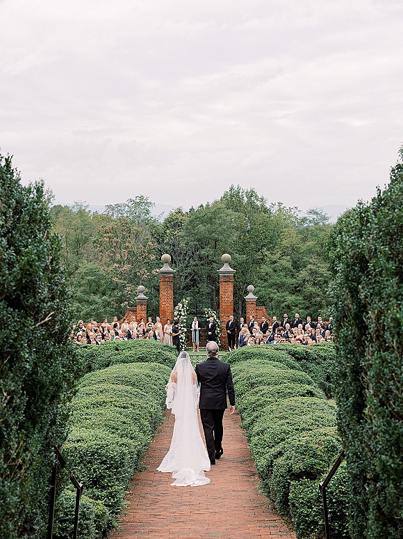 Wedding processional as bride walking down aisle with her father, cathedral veil trailing on a brick path between hedges and guests