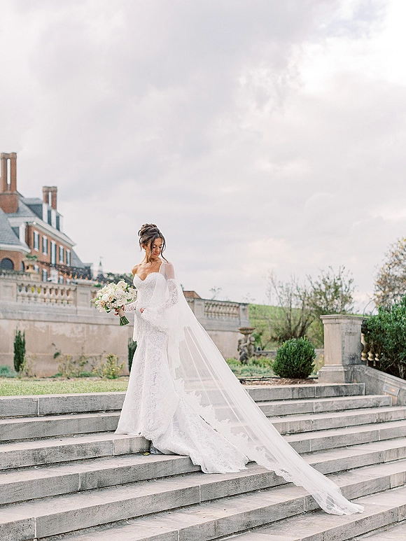 Bridal portrait of a bride on steps holding a white-and-blush bouquet, wearing a strapless lace gown and long veil by a historic terrace wall