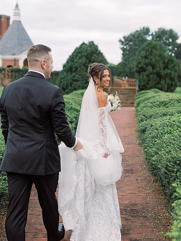 Couple portrait of bride and groom walking hand in hand on a brick garden path, groom lifting her train, bouquet and veil flowing