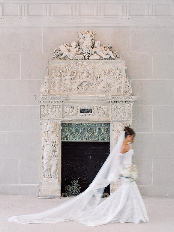 Bridal portrait of a bride in a wedding dress holding a white bouquet with a long veil trailing before an ornate stone fireplace indoors