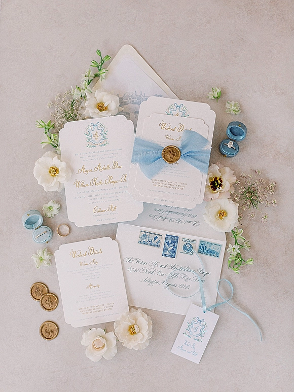 Invitation suite flat lay with blue and white wedding invitations, vellum ribbon, wax seal, stamps, rings and white flowers on stone surface