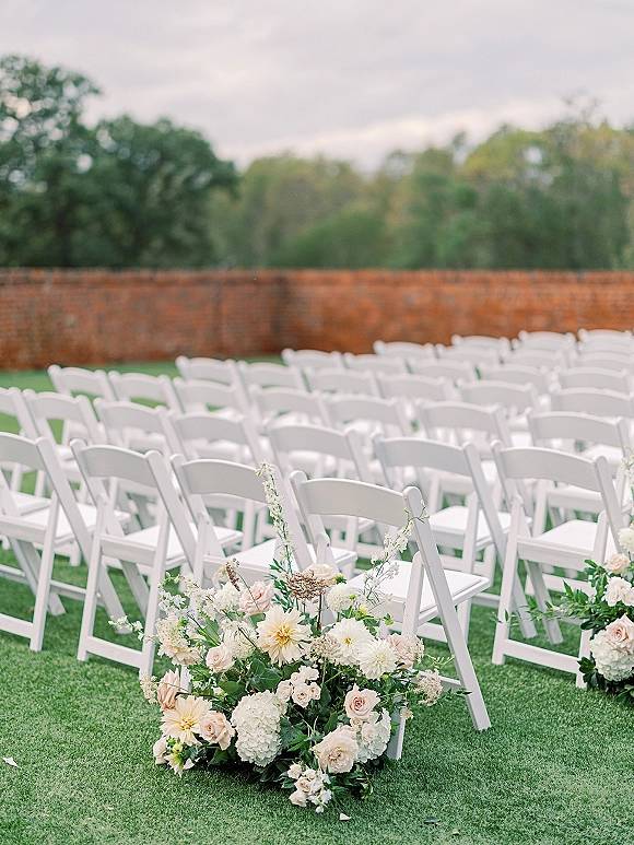 Ceremony seating with outdoor wedding ceremony chairs, white folding rows flanking an aisle lined with roses and greenery on a lawn by a brick wall