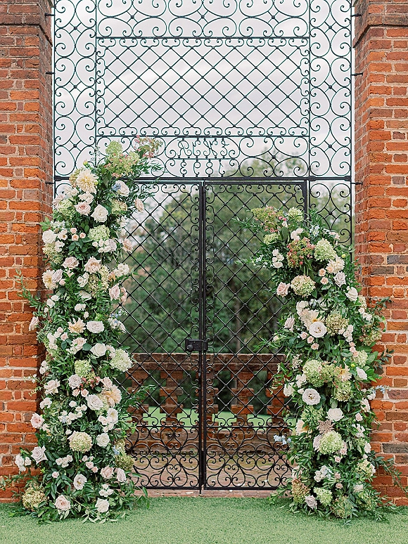 Wedding floral arch with an asymmetrical design of roses and hydrangeas, accented by greenery garland beside a wrought iron gate and brick wall