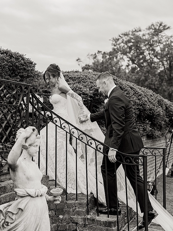 Couple portrait of bride and groom on stairs, holding hands as her cathedral veil flows down stone steps beside a brick wall and garden statue