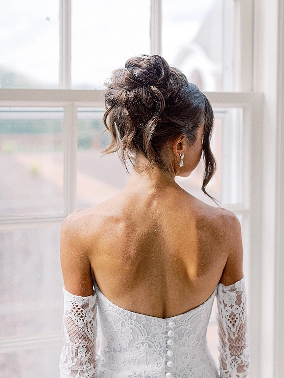 Bridal hair updo with a wedding updo hairstyle in a textured low bun, bride seen from back in lace off-the-shoulder dress by window light
