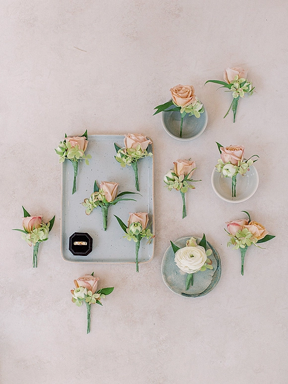 Wedding boutonnieres arranged in a flat lay beside a ring box and gold ring on a light neutral surface with pink rose accents