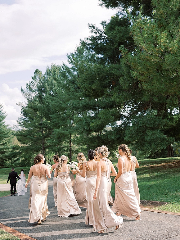Bridesmaids walking away in champagne bridesmaid dresses, holding bouquets and wearing heels on a tree-lined path under cloudy sky