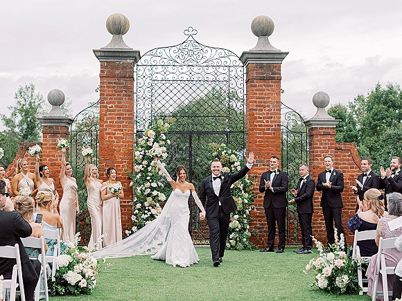 Wedding recessional as bride and groom cheer walking up the aisle, long veil trailing beneath a floral arch with white chairs and bouquets