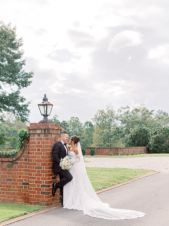 Couple portrait of bride leaning into groom, holding a white rose bouquet by an ivy-covered brick wall with lantern and cloudy sky
