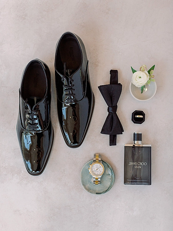 Groom details flat lay with black patent dress shoes, bow tie, boutonniere, wedding band, watch, and cologne on a light stone surface