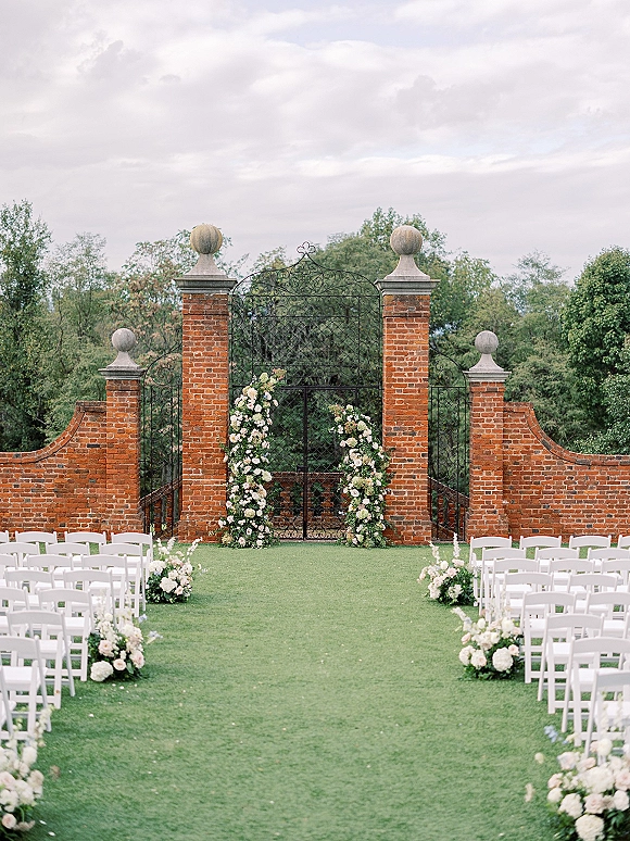 Ceremony setup for an outdoor wedding ceremony with white folding chairs lining a grassy aisle, floral arch and greenery by a wrought iron gate