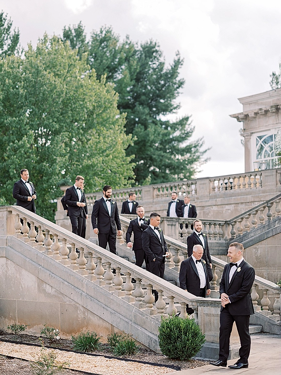 Groomsmen portraits of the groom and groomsmen walking down a stone staircase in black tuxedos, framed by terrace greenery under cloudy sky