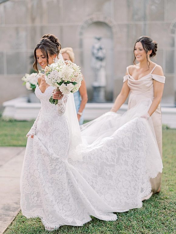 Bride portrait with bride holding bouquet of white and blush flowers in a lace long-sleeve gown and veil, by a stone courtyard fountain