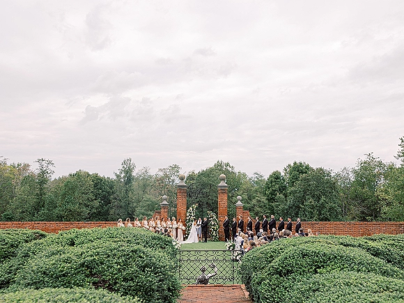 Wedding ceremony with bride and groom at altar beneath a floral arch, wedding party lined up by aisle runner near brick pillars and iron gate