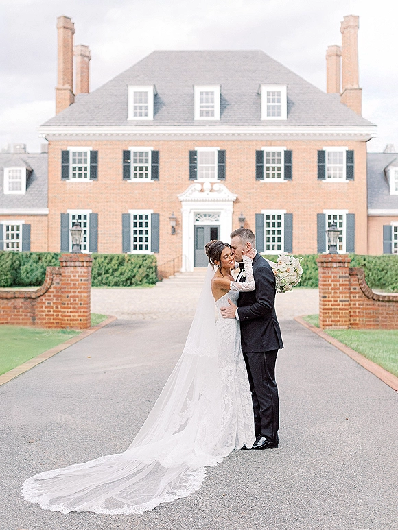 Wedding couple portrait of bride and groom embrace in front of a brick manor house, her long veil flowing as she holds a bouquet