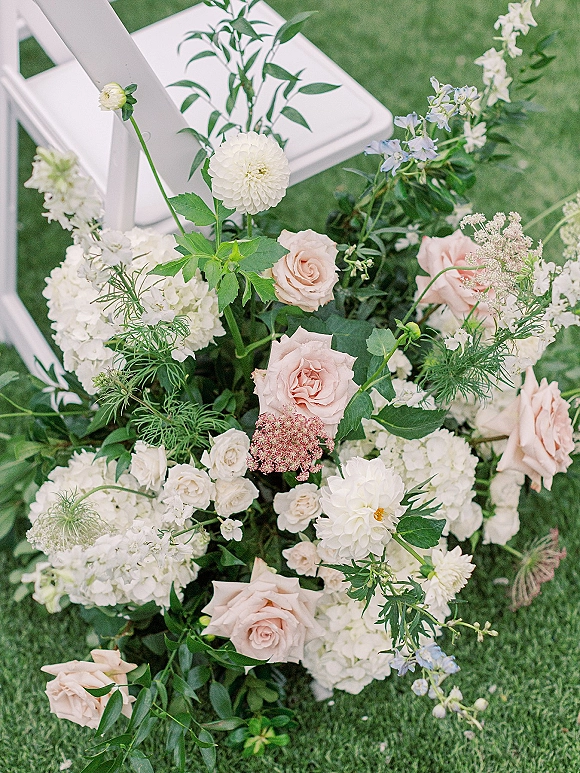 Ceremony aisle flowers in a low wedding aisle floral arrangement of blush roses, dahlias, hydrangea and greenery beside a white folding chair on grass lawn
