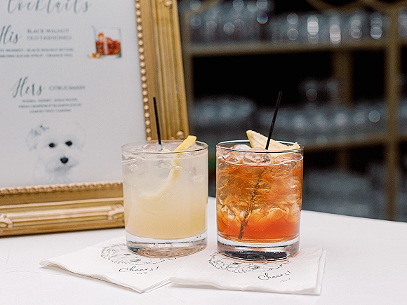 Signature wedding cocktails in rocks glasses with ice, black straws, and lemon peel garnish beside a gold-framed wedding cocktail bar menu sign
