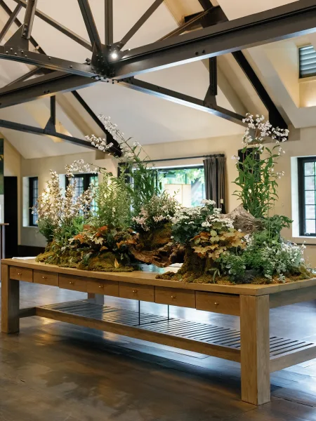 Reception centerpiece with greenery table runner, moss, driftwood, white flowers, and ferns on a wooden table in an indoor venue
