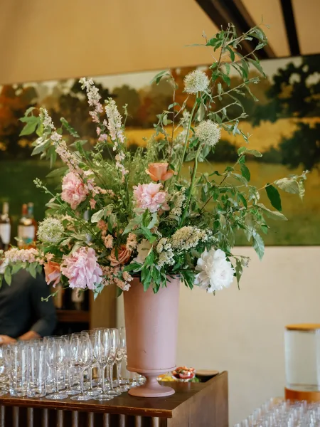Wedding floral centerpiece in a tall urn centerpiece with pink, peach, and white blooms and airy greenery on a bar counter by a mural wall