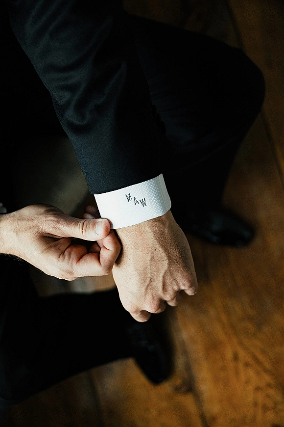 Groom getting ready in a dark suit jacket, showing groom suit detail with a handwritten note on his shirt cuff in an indoor room