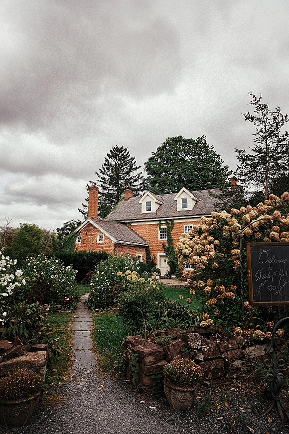 Wedding venue exterior with a wedding welcome sign on a chalkboard by a stone wall, leading down a hydrangea-lined garden path to a brick house